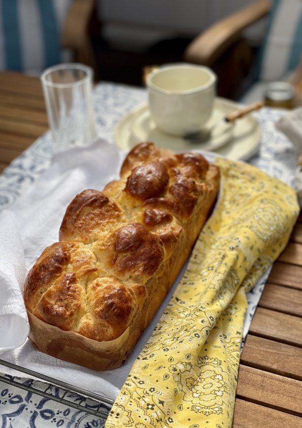 Brioche assado na cor dourada em diagonal sobre uma toalha de prato branca e amarela numa mesa de madeira ripada, com xícara de café